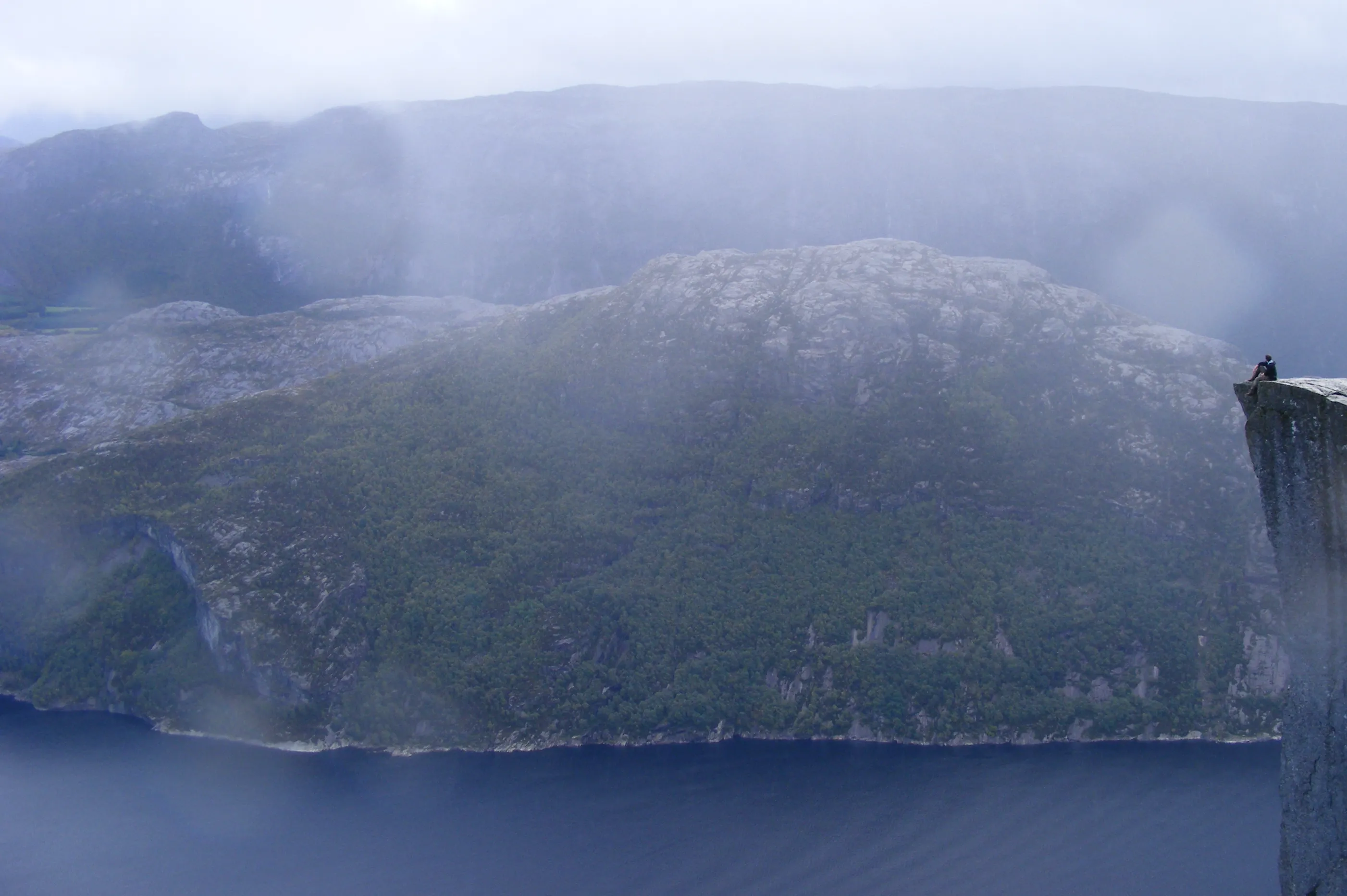 View of the Lyse Fjord with blue water at the bottom, blue sky at the top, and a very small me on the right side, sitting on the edge of the Preikestolen—a rocky massif that overlooks the fjord at 600 m.