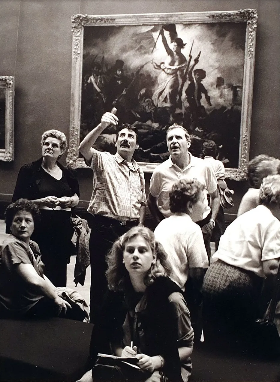 Schwarz-weiß Fotografie. Engagiert diskutierende Louvre-Besucher vor dem Gemälde „Die Freiheit führt das Volk“ von Eugène Delacroix, das unschafr im Hintergrund sitzt.