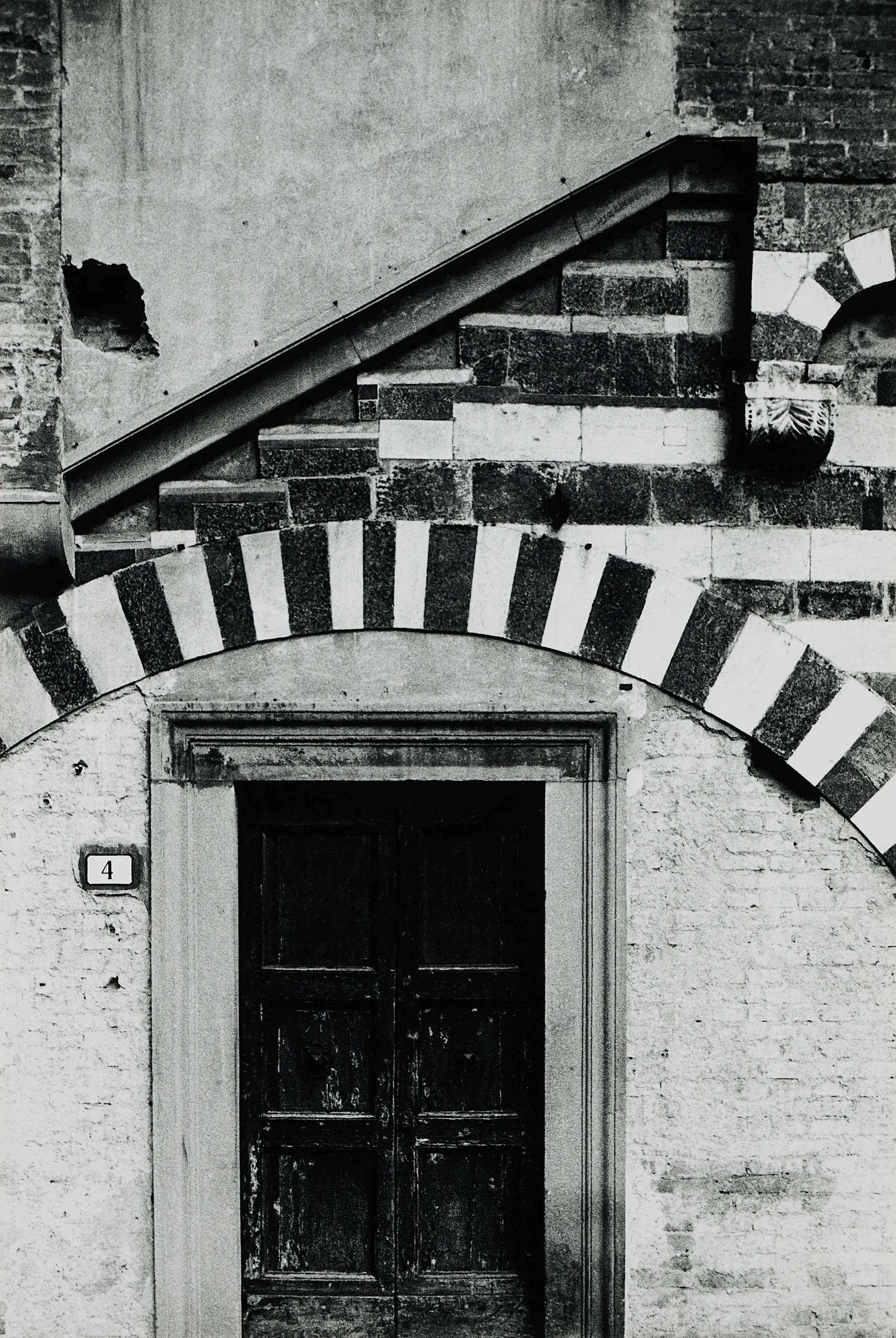 In der unteren Bildhälfte sieht man eine geschlossene Tür mit der Hausnummer 4. In der oberen eine ins Nichts führende Treppe. Niemand ist zu sehen.